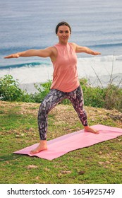 Young Woman Practicing Yoga, Standing In Virabhadrasana Pose. Outdoor Yoga On The Cliff. Warrior Pose. Work Out. Healthy Lifestyle. Ocean In The Background. Yoga Retreat, Bali.