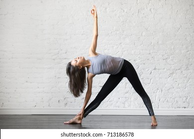 Young woman practicing yoga, standing in Utthita Trikonasana exercise, extended triangle pose, working out, wearing sportswear, gray tank top, black pants, indoor full length, studio background 