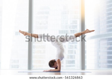 Young woman practicing yoga, standing in handstand exercise, variation of Pincha Mayurasana pose, working out, wearing white sportswear, indoor full length, floor window with city view. Copy space 