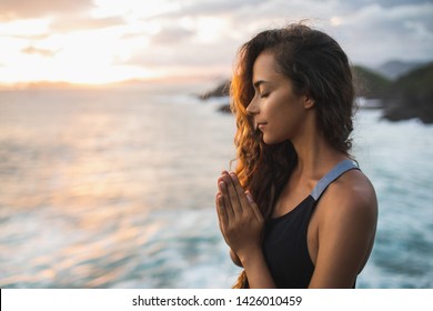 Young woman praying and meditating alone at sunset with beautiful ocean and mountain view. Self-analysis and soul-searching. Spiritual and emotional concept. Introspection and soul healing.