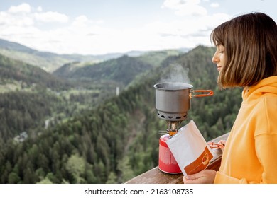 Young woman prepares freeze-dried food for hiking in airtight package, boiling water with gas burner, while traveling in mountains on summertime. Food concept during hiking