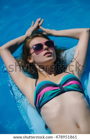 Young woman with pretty face beautiful abdomen and breast in colorful bikini and sunglasses tanning and relaxing on air bed in blue water of pool in summer