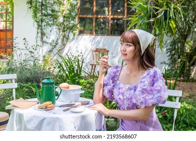 Young Woman In A Princess Dress Sits Drinking Tea On A Vintage Table With Cookies And Books In A Backyard With Plants And Flowers.