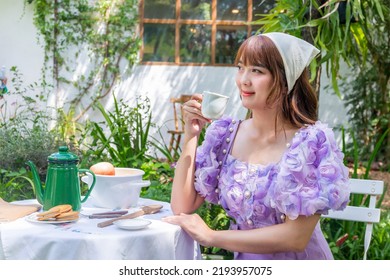 Young Woman In A Princess Dress Sits Drinking Tea On A Vintage Table With Cookies And Books In A Backyard With Plants And Flowers.