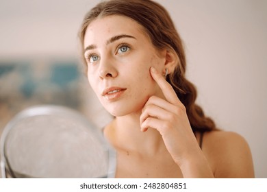 Young woman with problem skin looking into mirror. Concept of acne therapy, scars, inflammation on face, problem skin. Beauty care.