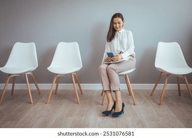 A young woman in professional attire sits in a waiting room using her smartphone. The mood is calm and focused, suggesting a business environment or job interview preparation. - Powered by Shutterstock