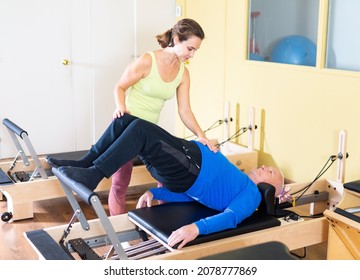 Young woman professional pilates instructor helping aged man doing exercises on reformer. Wellness concept for elderly