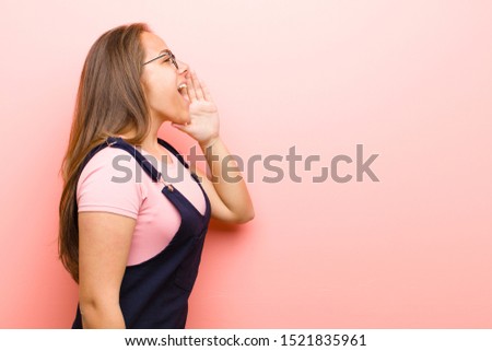 young  woman profile view, looking happy and excited, shouting and calling to copy space on the side against pink background
