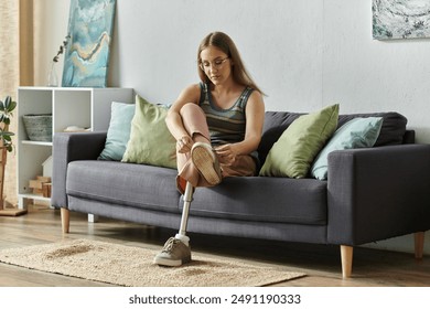 A young woman with a prosthetic leg sits on a couch in her living room, tying her shoe. - Powered by Shutterstock