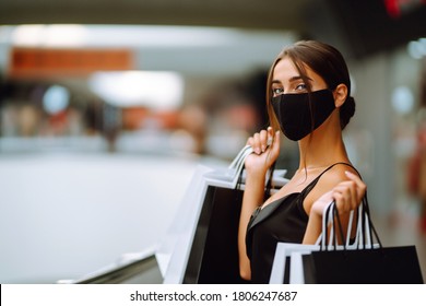 Young Woman In Protective Black Medical Mask On Her Face With Shopping Bags In The Mall. Purchases, Black Friday, Discounts, Sale Concept. Covid-2019.