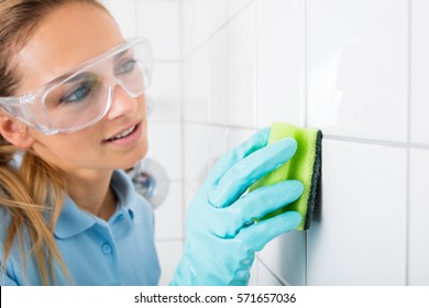 Young Woman With Protective Eyewear Cleaning The White Tile Of The Wall Using Sponge