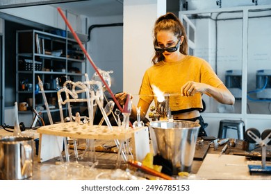 A young woman in protective eyewear uses a torch to heat glass in a studio workshop, surrounded by tools and materials for glassblowing. - Powered by Shutterstock