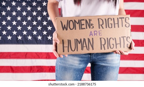 Young Woman Protester Holds Cardboard With Womens Rights Are Human Rights Sign Against USA Flag On Background. Feminist Power. Equal Opportunity Womens Rights Freedom