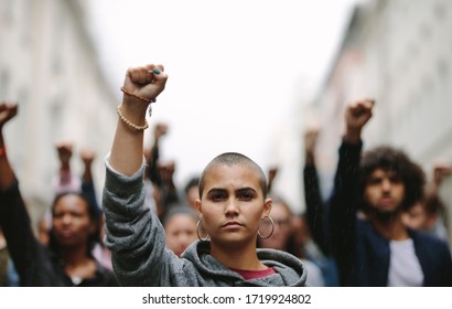 Young woman protesting on the street with her fist raised in air. Group of protesters on the road with their arms raised. - Shutterstock ID 1719924802