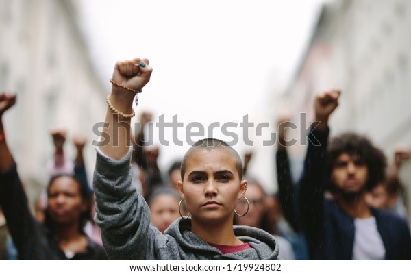 Young\
woman protesting on the street with her fist raised in air. Group\
of protesters on the road with their arms\
raised.