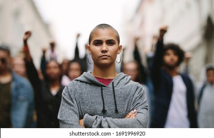 Young woman protestor standing with her arms crossed and group of people in background on the city street. Activists demonstrating in the city. - Shutterstock ID 1691996920