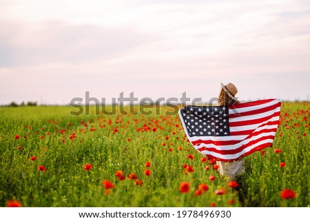 Young woman proudly hold waving american USA flag in in the poppy field. Patriot raise national american flag against the blue sky. Independence Day, 4th July.
