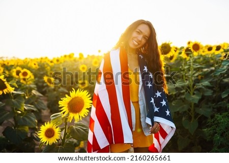 Young woman proudly hold waving american USA flag in in the sunflower field. Patriot raise national american flag at sunset. Independence Day, 4th July.