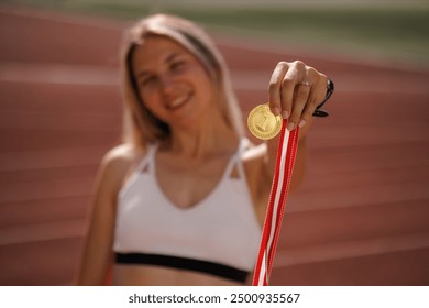 Young woman proudly holding gold medal with red and white ribbons standing on athletics field. Winner in athletics competition - Powered by Shutterstock