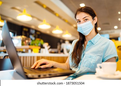 Young Woman In A Public Place Working On Laptop, Wearing Protective Face Mask Indoors.Online Training Education And Freelance Work.Studying Remotely.Outsourcing. Coronavirus Pandemic Concept