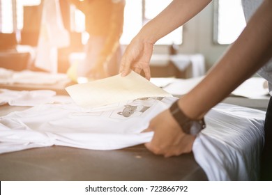 Young Woman Pull Out Paper From Waterproof Film On Fabric. Worker Working On Manual Screen Printing On T-shirt At Her Shop.