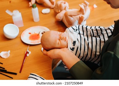 Young Woman Puppeteer Holding In Her Hands Small Cute Handmade Reborn Baby Doll At Workplace, Indoors. Craft Making Dolls, Hobby Reborning. View From Above.