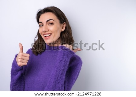Young woman in a purple soft cozy sweater on a background of cute smiling cheerfully, in high spirits, points a finger to the right to an empty space	