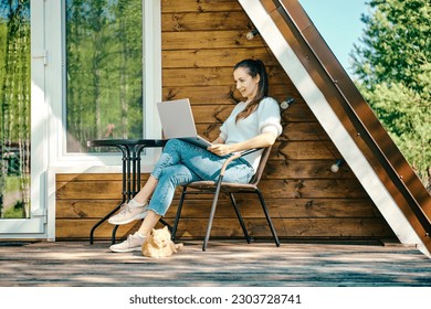 Young woman put laptop on her knees while resting on terrace of wooden a-frame cabin