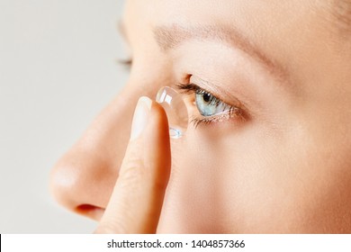 Young woman puts contact lens in her eye over white background. Eyewear, eyesight and vision, eye care and health, ophthalmology and optometry concept. Selective focus, close up