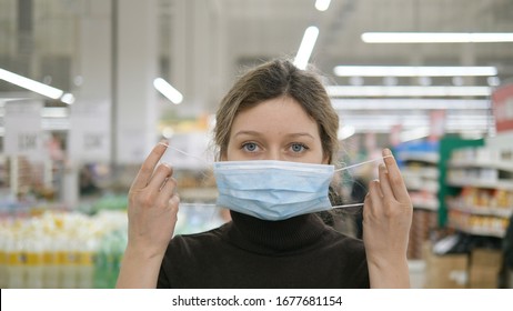 A Young Woman Puts On A Medical Mask In A Grocery Supermarket Close-up And Looks At The Camera, Protection From Coronavirus, Shopping In A Store To Wait Out The Quarantine.