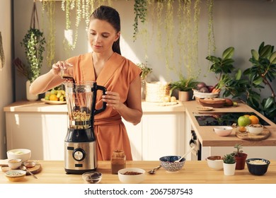 Young woman putting all the ingredients for homemade smoothie or dessert into electric blender while cooking in the kitchen - Powered by Shutterstock