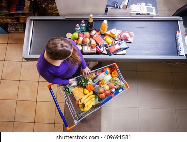 Young woman putting goods from shopping cart on counter for checkout in supermarket