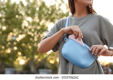 Young woman putting smartphone in stylish bum bag outdoors, closeup