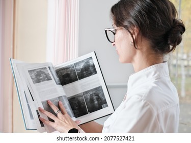 Young Woman Radiologist Referring To A Book To Find Information On Diagnostic Issues. Woman Wearing Medical Uniform And Glasses Holding A Book. Side View