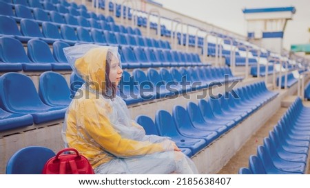 Young woman in raincoat sitting on stadium bleachers alone in rainy weather. Female spectator in wet waterproof coat at street stadium during competitions