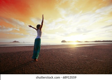 Junge Frau mit hohen Händen, die auf nassem Sand stehen und einen roten Himmel sehen