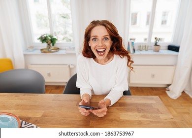 Young woman reacting in amazement and delight staring wide eyed at the camera with an animated expression as she sits at a table at home with her mobile phone - Shutterstock ID 1863753967