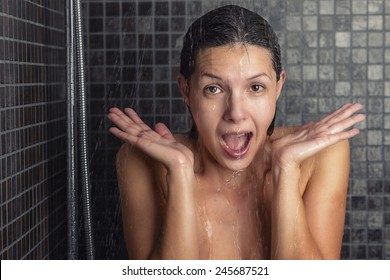 Young woman reacting in shock to hot or cold shower water as she stands under the shower head washing her hair looking at the camera with her hands raised and mouth open