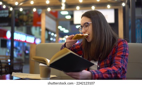 A young woman is reading a book in a cozy cafe. A woman sits at a table and eats pizza in the food court. Girl in a red shirt with glasses. - Shutterstock ID 2242123107