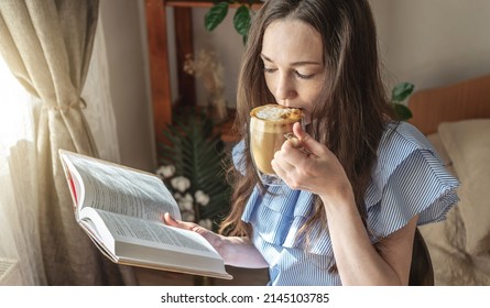 Young woman is reading a book and drinking coffee sitting by the window. Concept of morning enjoyment, leisure, pleasant pastime and relaxation.