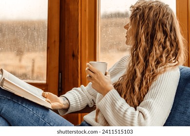 Young woman reading book and drinking coffee or cocoa at indoor terrace. Beautiful female with cup of tea passionate about reading exciting novel while sitting in armchair and looking out window rain. - Powered by Shutterstock