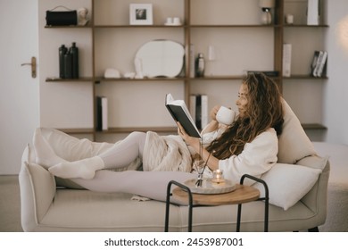 Young woman reading book and drinking tea, relaxing on sofa in a cozy living room. - Powered by Shutterstock