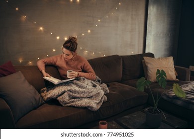 Young Woman Reading Book And Drinking Coffee On Sofa In Hygge House. Caucasian Female Relaxing In Cozy Living Room And Reading A Book.