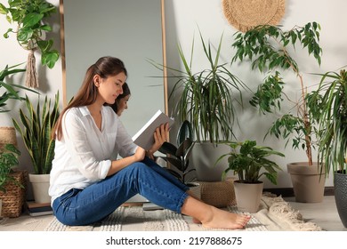 Young Woman Reading Book Near Mirror And Different Houseplants On Floor In Room