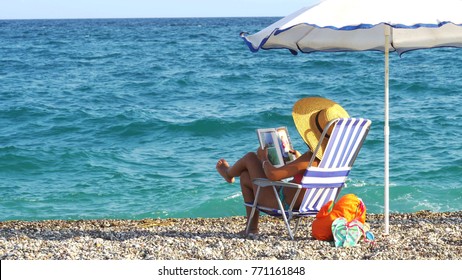 Young Woman Reading A Book On Beach Drinking Hot Tea Or Coffee From Thermos