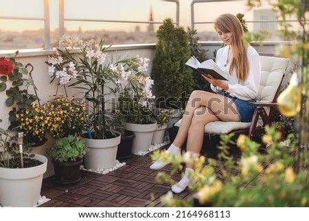 Young woman reading a book on urban rooftop garden. Girl with paper storybook sitting in chaise longue on cozy terrace with flowers, plants and city view. Female enjoying life on cozy green balcony.