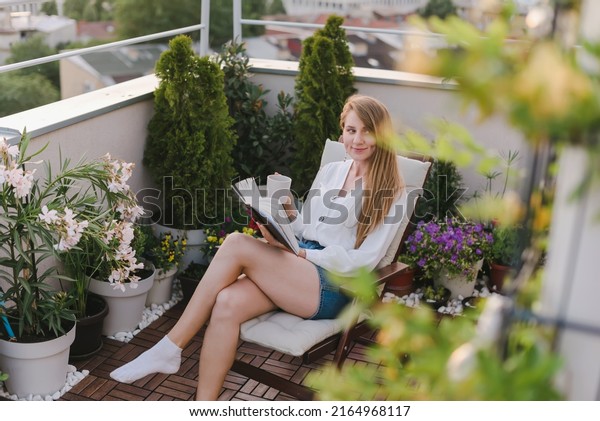 Young woman reading a book on urban rooftop\
garden. Girl sitting in chaise lounge and relaxing with cup of tee\
or coffee and paper book. Female enjoying life on cozy terrace with\
flowers and plants.