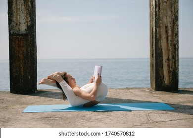 Young woman reading a book while practicing yoga at seaside.  Woman doing yoga pose with legs behind her head
