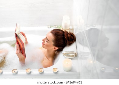 Young Woman Reading Book While Lying In The Bath With Foam And Candles