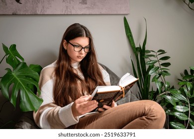 Young woman reading a book while sitting indoors, surrounded by green plants in a peaceful setting - Powered by Shutterstock